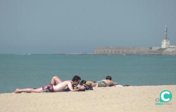 Unos jóvenes toman el sol sobre la arena con el castillo de San Sebastián al fondo