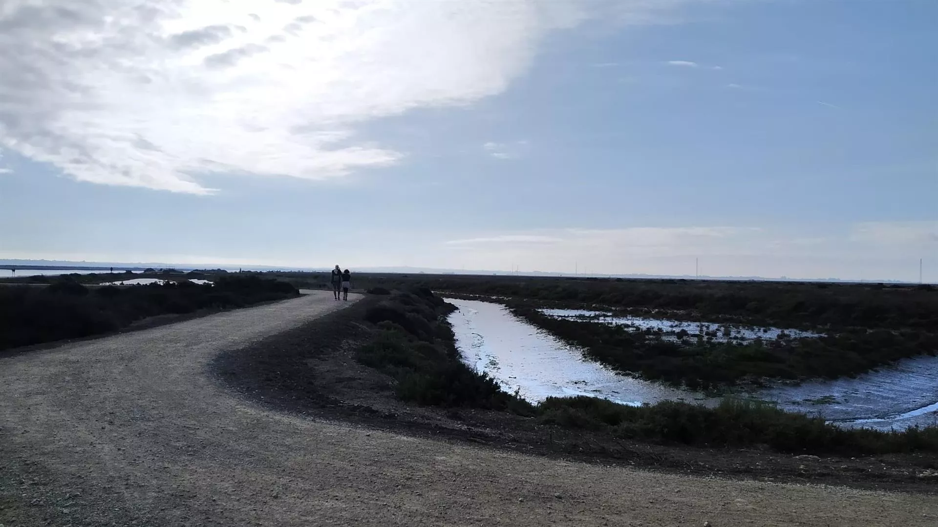 Viandantes caminan por un paseo junto a unos esteros de la Bahía de Cádiz