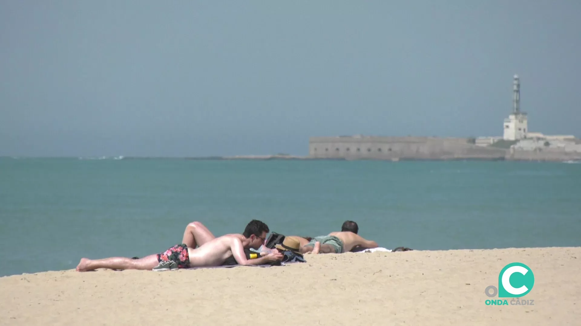 Unos jóvenes toman el sol sobre la arena con el castillo de San Sebastián al fondo